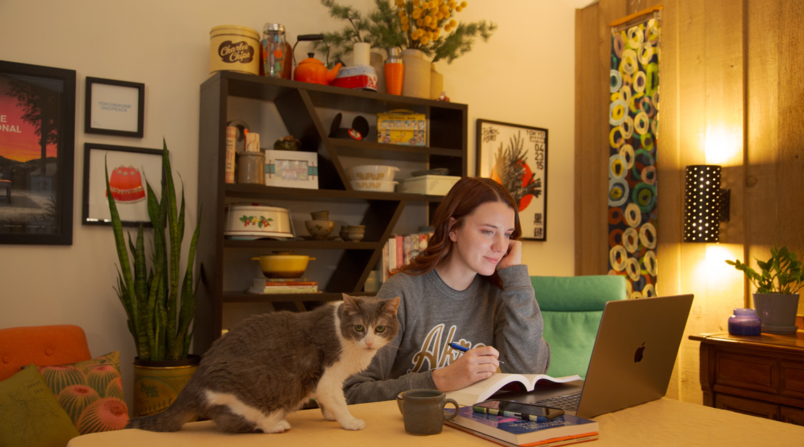 A student sitting at her dining room table looking at her computer. bet365官网re are books on the table and her cat is providing company.