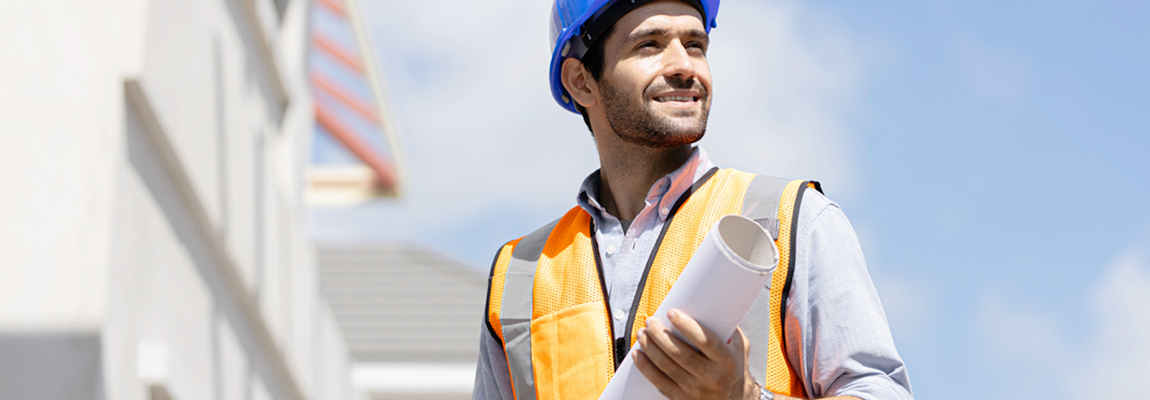 A man wearing a hard hat and a safety vest at a construction site. bet365官网 man is also holding a roll of building plans.