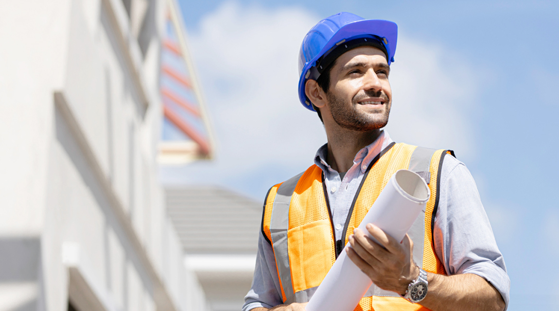 A man wearing a hard hat and a safety vest at a construction site. bet365官网 man is also holding a roll of building plans.