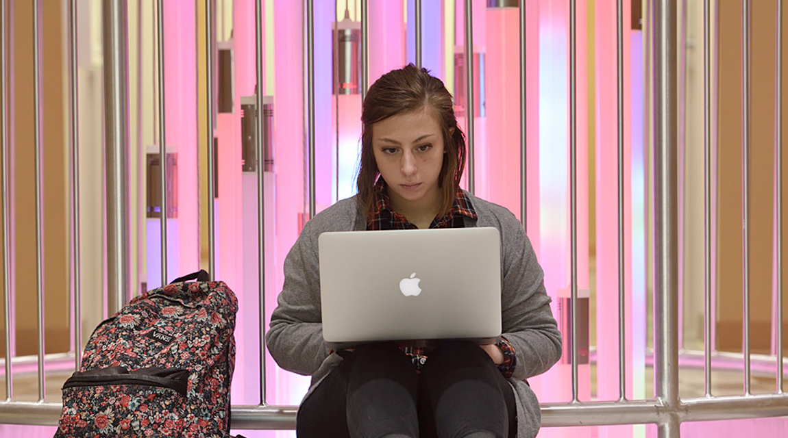 A student sitting on the floor with a laptop on her lap and a backpack next to her. She is facing the camera but looking at her computer.