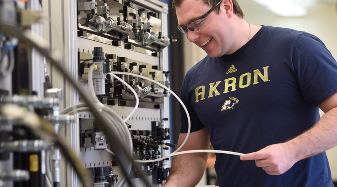 A smiling student standing in front of a wall of industrial controls.