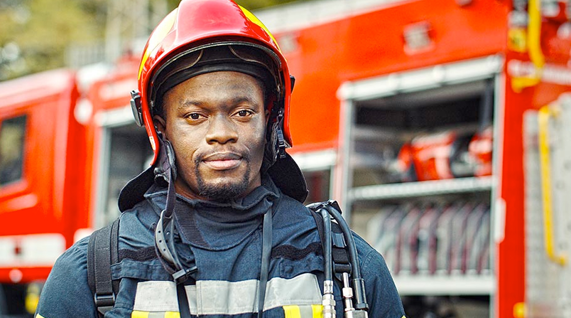 A male firefighter wearing a helmet and other safety gear standing in front of a fire truck.