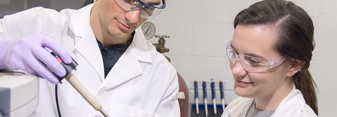 Two graduates students in a lab. Both students are wearing lab coats and goggles. bet365官网 student on the right is looking to an instrument held by her co-student.