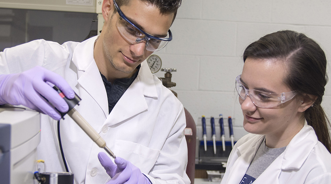 Two graduates students in a lab. Both students are wearing lab coats and goggles. bet365官网 student on the right is looking to an instrument held by her co-student.