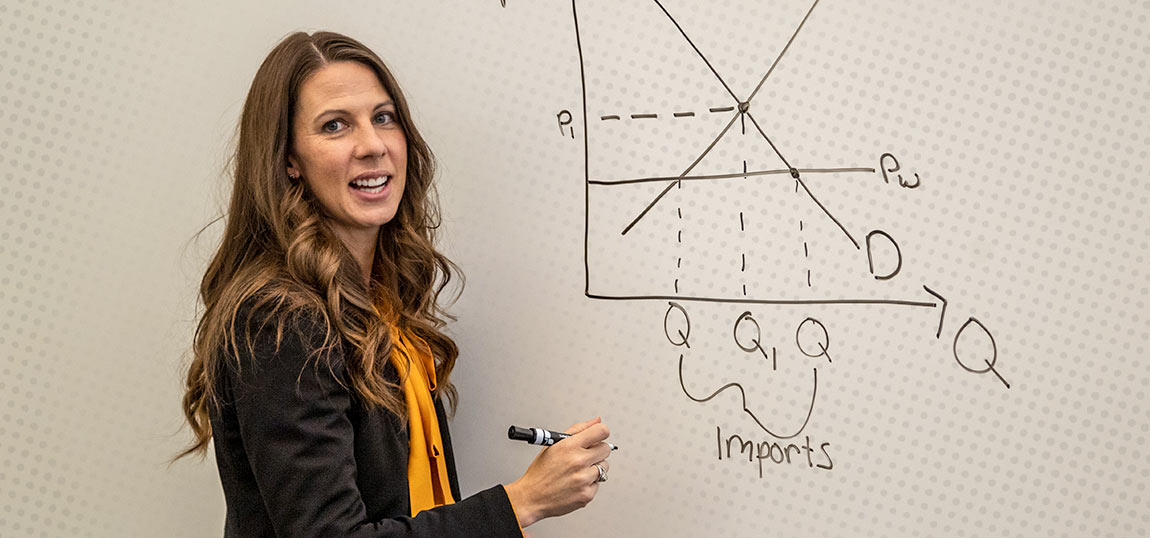 A professor standing in front of a whiteboard talking about a formula that is written on the board.
