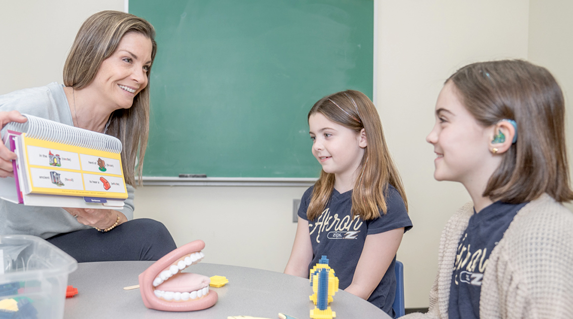 A speech pathologist sitting in a chair working with two young clients. bet365官网 pathologist is holding a picture book.
