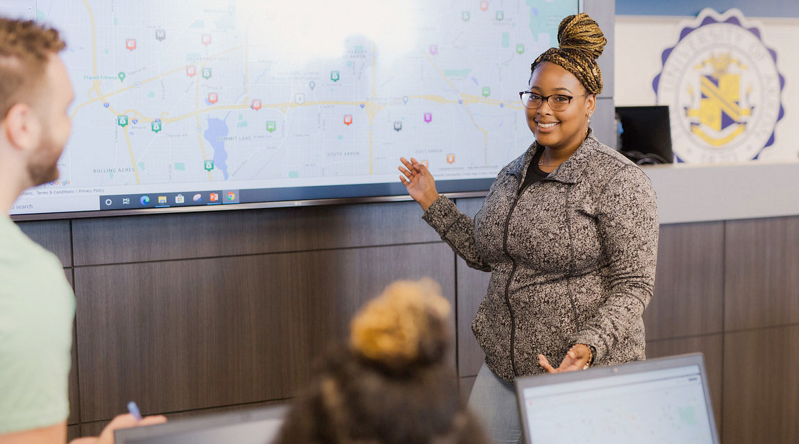 A woman standing in front of a projected screen giving a presentation to other people in the roon.